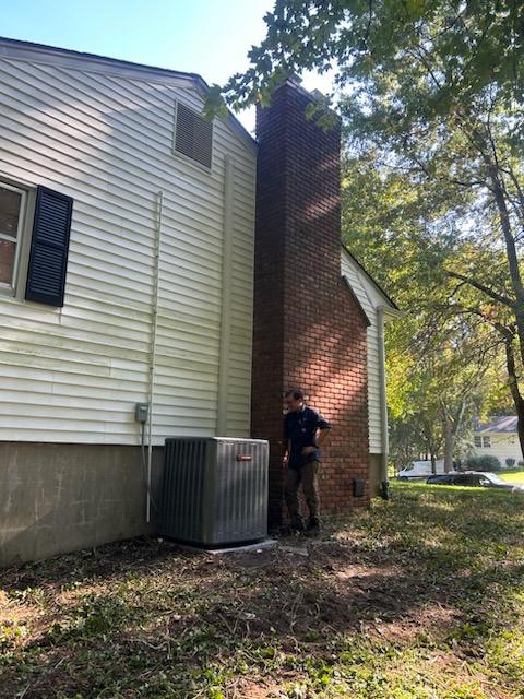 Technician standing beside an outdoor HVAC condenser unit installed next to a home with vinyl siding and a brick chimney