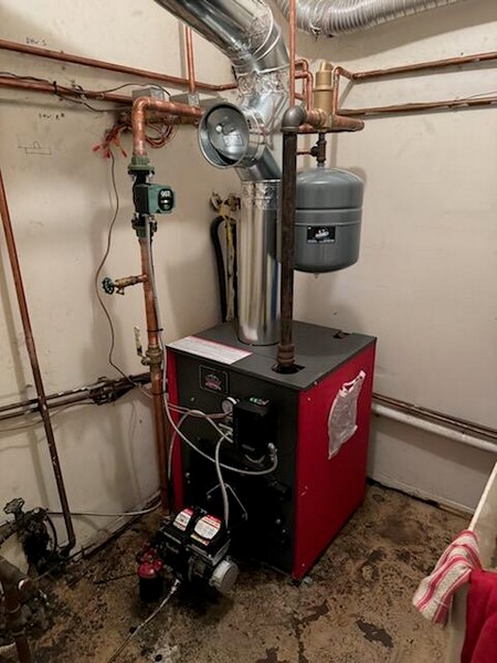 Residential boiler system installed in a basement, featuring a red boiler unit with copper piping, circulation pump, expansion tank, and metal venting.