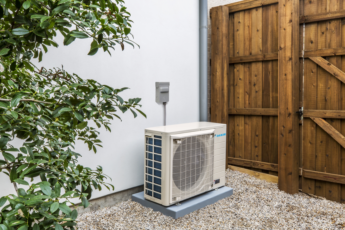 Outdoor heat pump unit installed beside a home on a gravel base near a wooden fence
