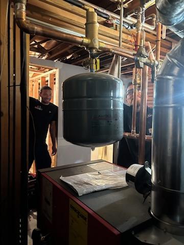HVAC technicians working on a residential boiler system with expansion tank and piping in a basement.