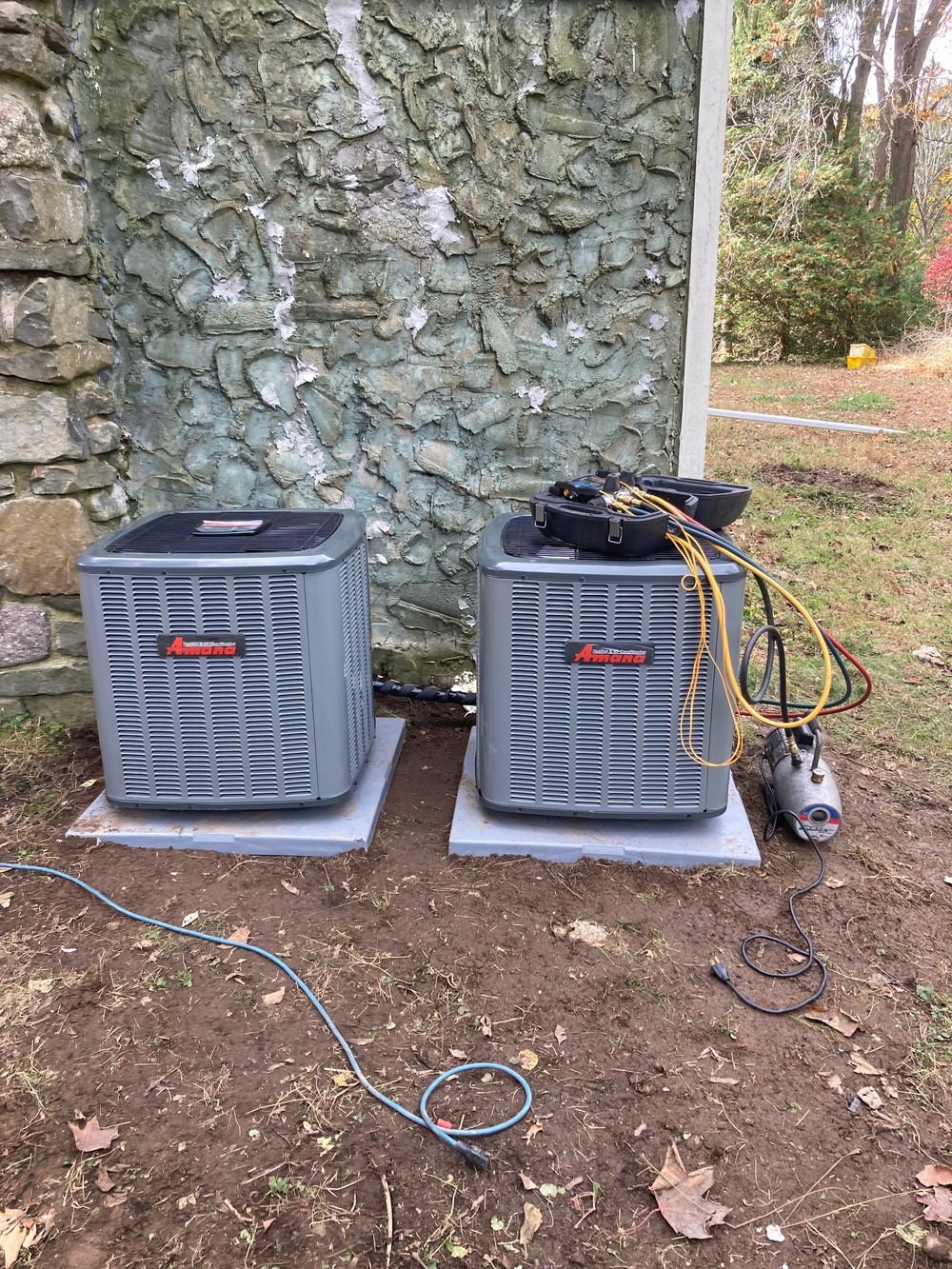 Outdoor air conditioning units installed on concrete pads beside a stone wall, with service tools and hoses visible nearby
